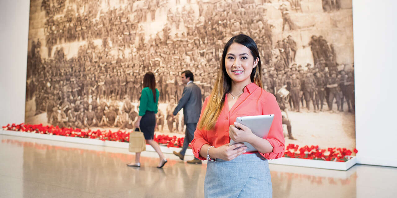 A woman standing in a gallery with a tablet in front of a large picture of soldiers and poppies.