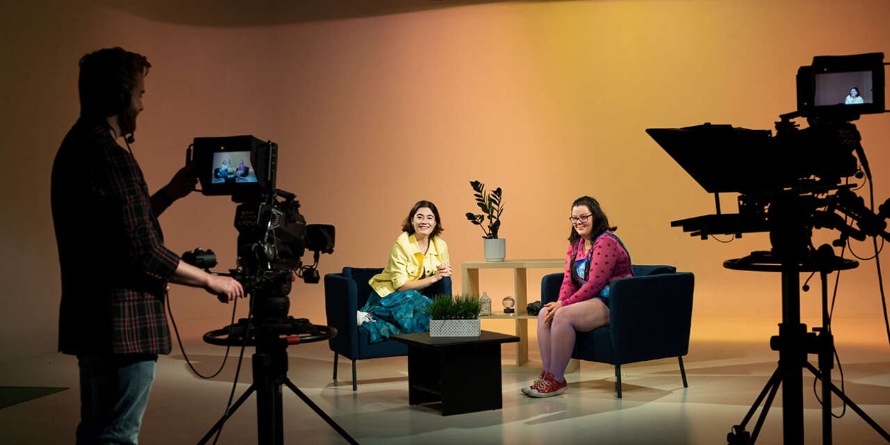 Two students sitting on film set in chairs while another student films.