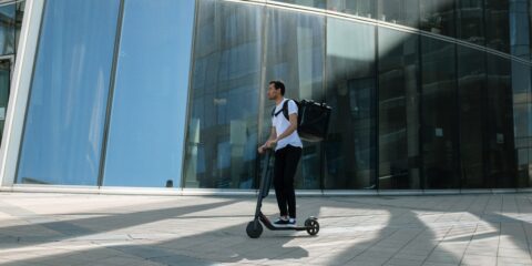 A person rides an e-scooter outside past a building.
