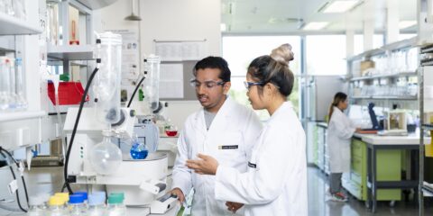 Two students in a laboratory wearing a laboratory coat and safety glasses.