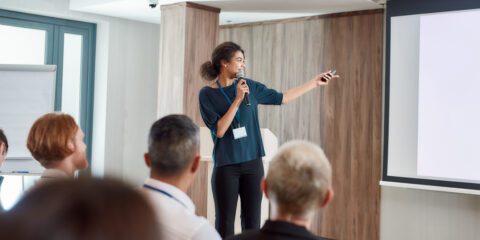 A woman presents at the front of a room pointing to the projector screen with a handheld laser pointer.