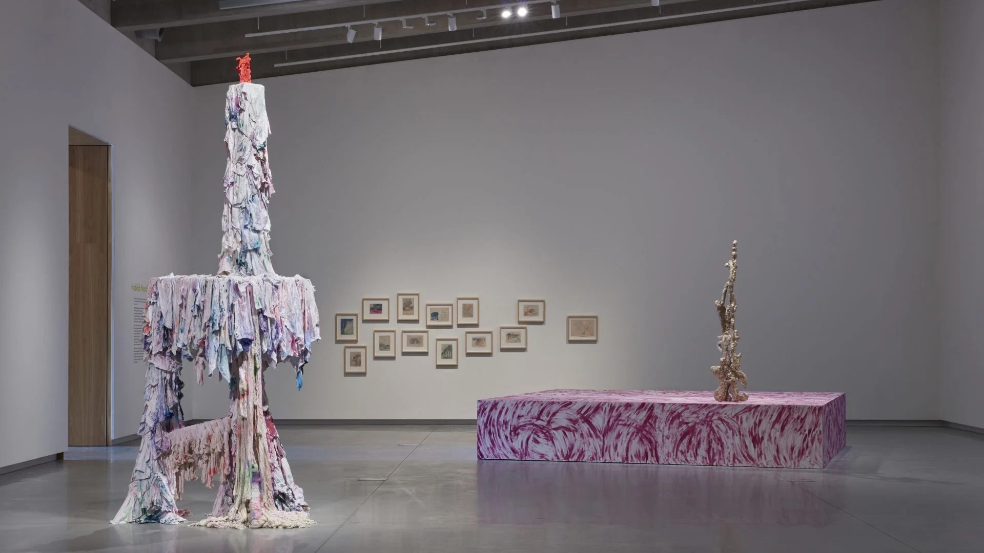 Two sculptures on a gallery floor infront of curated framed artworks on a gallery wall.