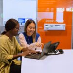 A library staff member helps a student sitting at a desk with their laptop