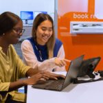 A library staff member helps a student sitting at a desk with their laptop