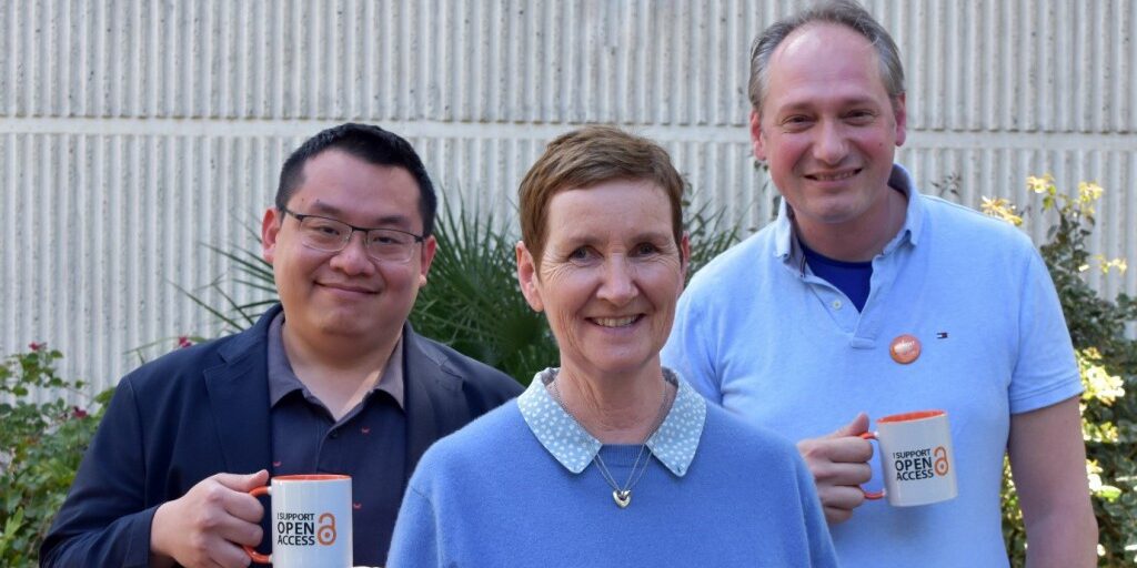 Dr Hendra Gunosewoyo, Catherine Clark (University Librarian) and Dr Alan Payne stand outside in front of bushes holding mugs that say 