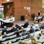 Rows of people sit and listen to University librarian Catherine Clark who is speaking at the lecture podium. She wears a green jacket and glasses.