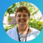 Jill wears a white blouse and Curtin lanyard. She has orange hair is standing outside smiling at the camera in front of trees and dappled light.