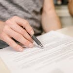 Close-up of a person's hand signing an important legal document with a pen indoors.