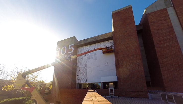 artist works on black and white mural on the side of Robertson Library from cherry picker