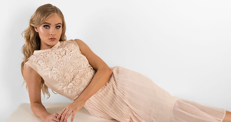Model wears long pleated dress with lace embroidery.