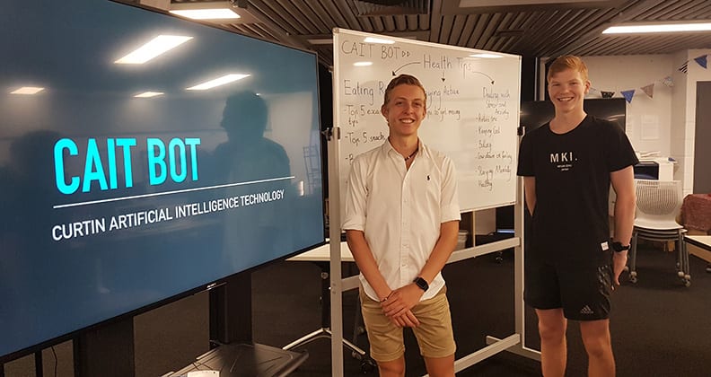 Two male high school students stand in front of a white board and computer screen with chatbot logo.