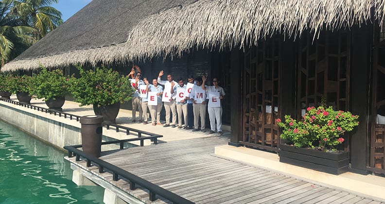 Eight staff hold a welcome sign beneath a large bungalow beside the water's edge.
