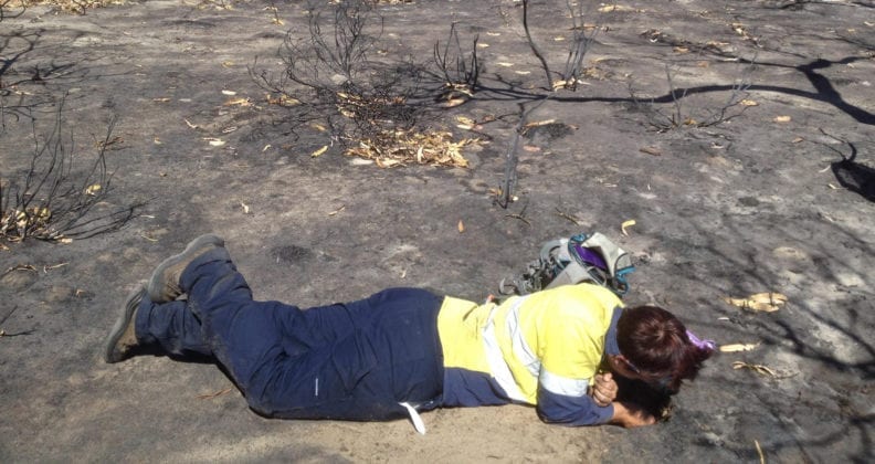 Leanda lays on the burned earth in bushland, examining a trapdoor burrow. 