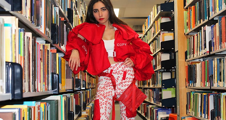 A yong girl leans against a library bookshelf wearing a bright, structured red jacket and white pants with the word 'Copy' printed over them. 
