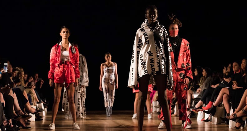 Five models stand on a dark catwalk wearing red, white and black clothing. 