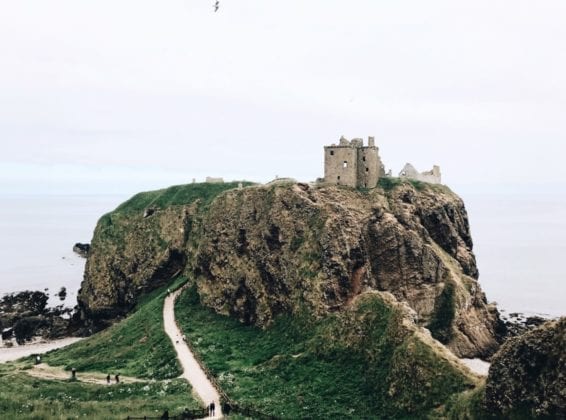 An old castle sits atop a large, rocky, green hill at the edge of the ocean.
