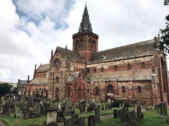 A red brick, old spired church with gravestones in the foreground.
