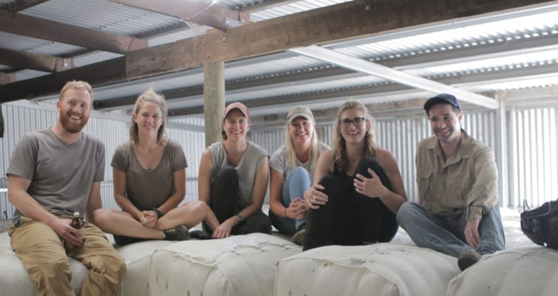Bignell and five other people sit on wool bales together in a large shed.