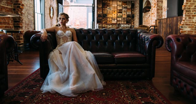 Bride wears a lace bodice and tulle gown on a dark leather couch.