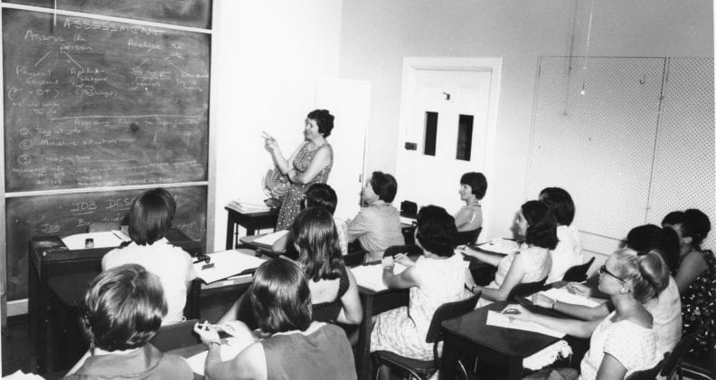 A black and white photograph of female teacher instructing a room full of students. The teacher points at the blackboard, which details information about how to assess a patient.