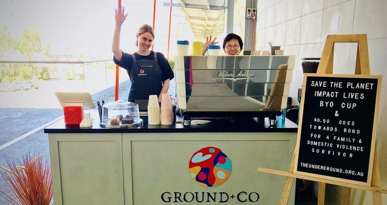 Two women wave happily from behind a coffee cart. 