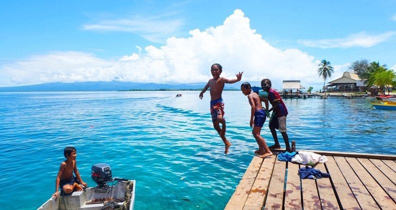 A young boy jumps off a jetty into aqua waters, while his three friends look on. 