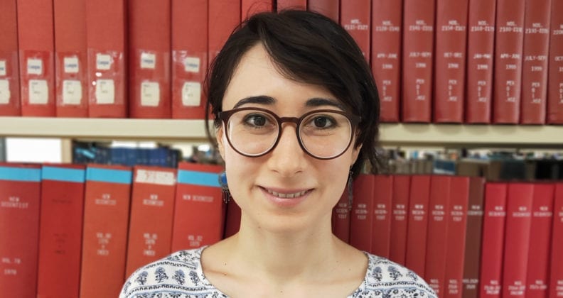Landscape photo of Anie Agius in front of red bound library books.