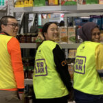 Three students stand in a food bank volunteering on the food shelves wearing food bank hi-vis.