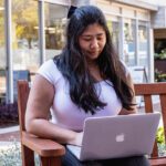 Student sitting on a bench working on a silver laptop.