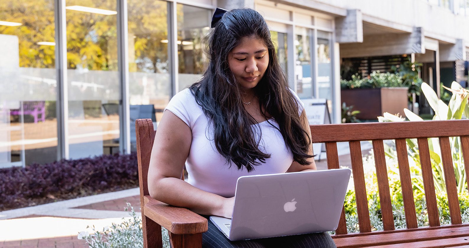 Student sitting on a bench working on a silver laptop.