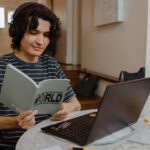 Student sitting at a round table reading a grey book with a black laptop in front of them.
