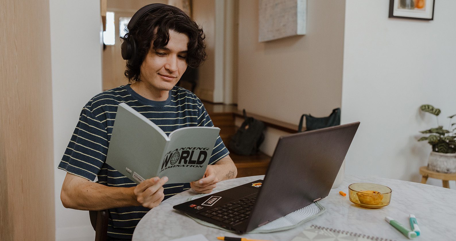 Student sitting at a round table reading a grey book with a black laptop in front of them.