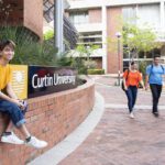 Male sitting on a retainer wall next to a Curtin university sign at Curtin campus, with people walking in the background