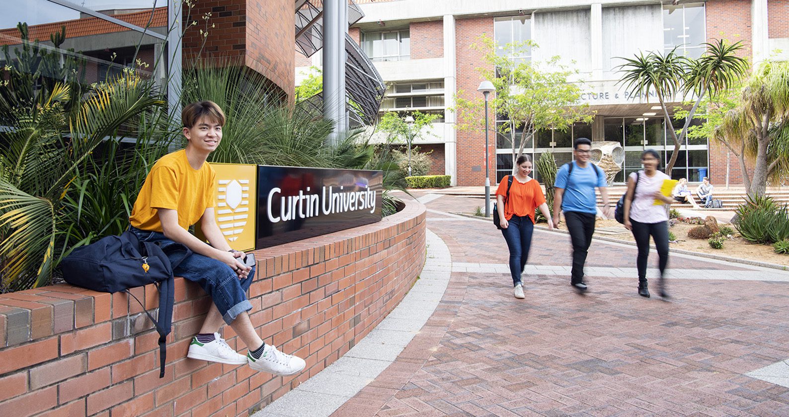 Male sitting on a retainer wall next to a Curtin university sign at Curtin campus, with people walking in the background