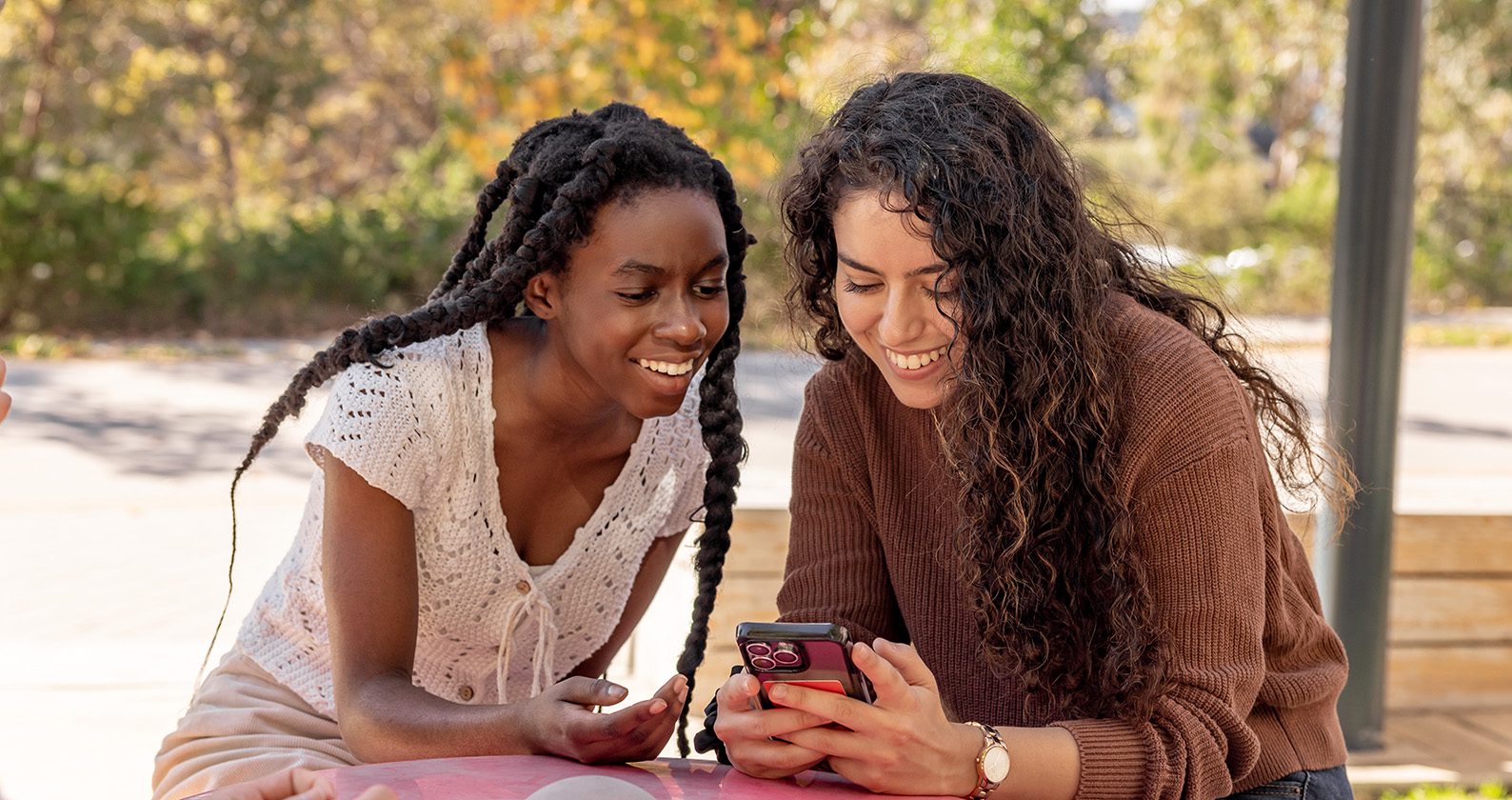 Two students sitting on a table, looking at the screen of one phone.