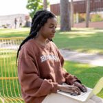 A female student sitting on a yellow chair, outside in a grassy area, and using her laptop.