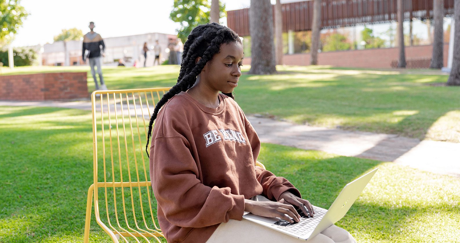 A female student sitting on a yellow chair, outside in a grassy area, and using her laptop.