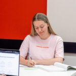 A student writes in her notebook with a textbook open.