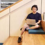 A male student sitting on some wooden steps looking at a laptop while wearing headphones