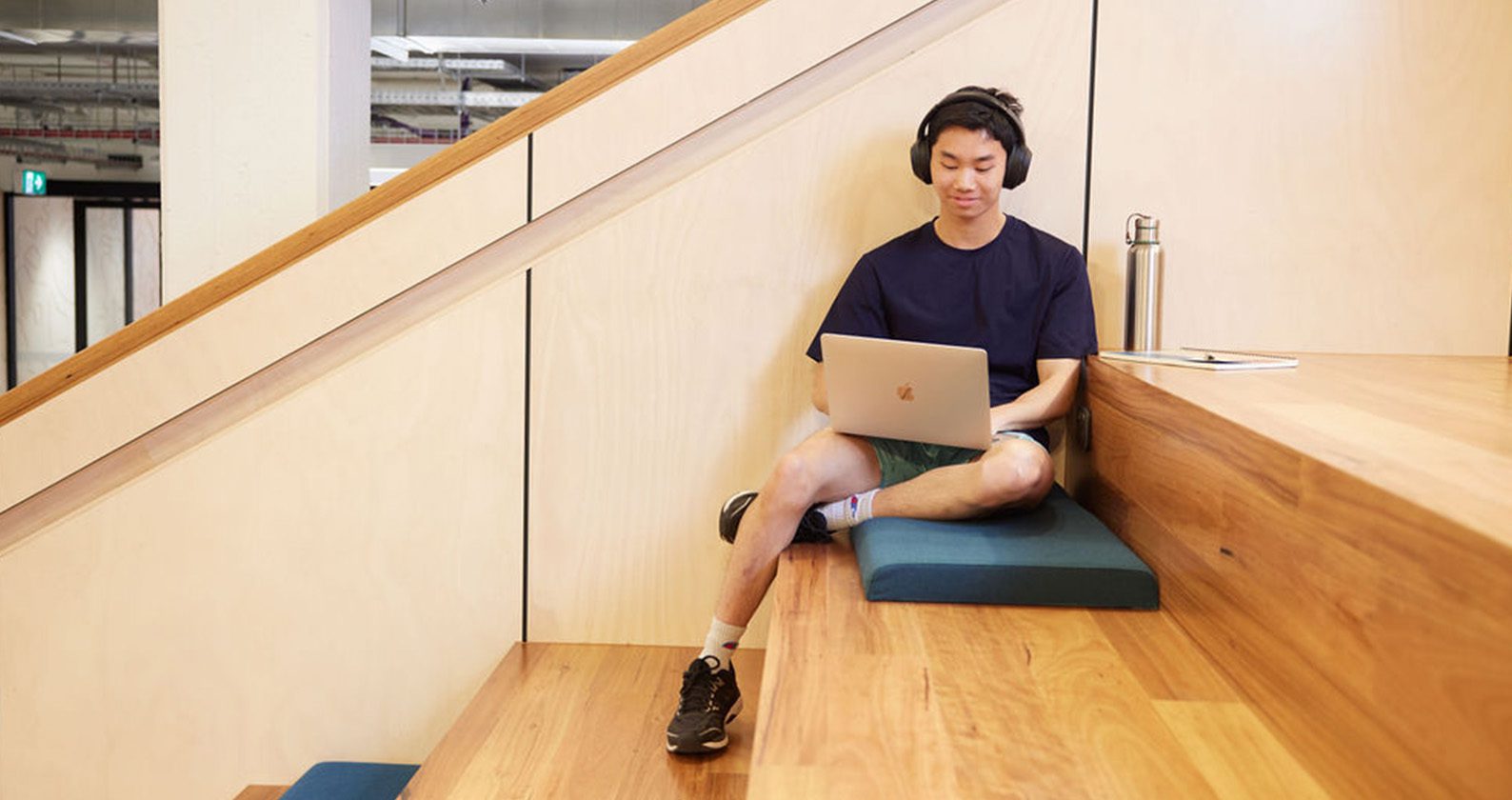 A male student sitting on some wooden steps looking at a laptop while wearing headphones