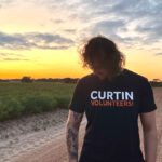 A Curtin Volunteer looking towards the grounds wearing the Curtin Volunteers shirt. He stands against a sunset and dirt road.