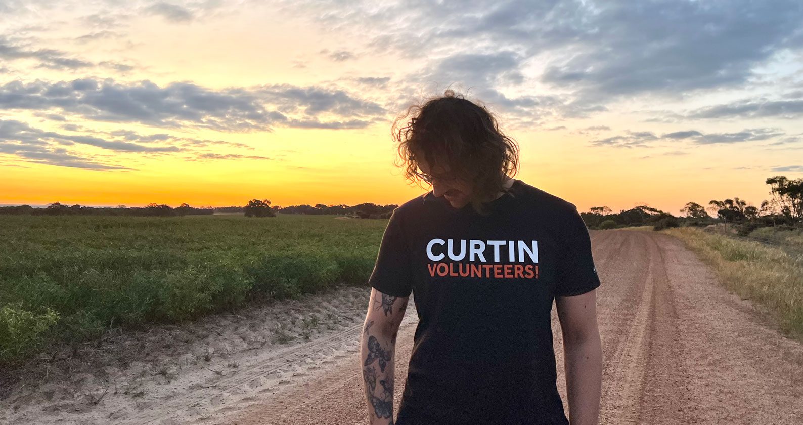 A Curtin Volunteer looking towards the grounds wearing the Curtin Volunteers shirt. He stands against a sunset and dirt road.