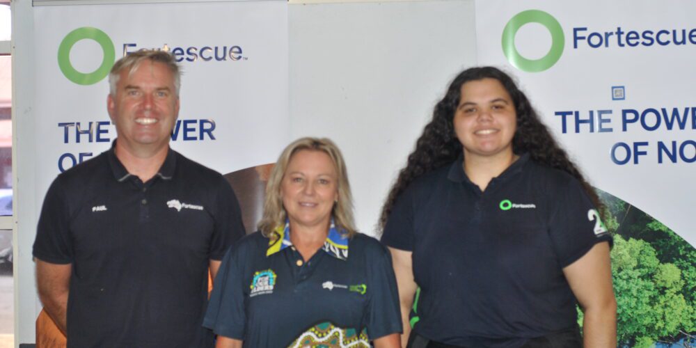 Three Fortescue Metals staff members wearing company polos standing in front of pull up banners. 