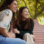 two young students laughing on a bench together