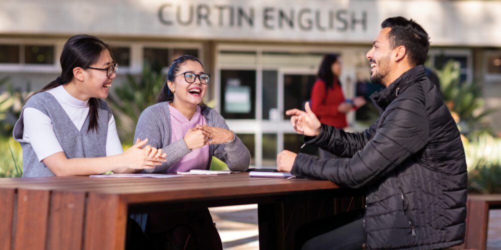 Three Curtin English students sitting at a picnic bench outside the Curtin English building, chatting and laughing.