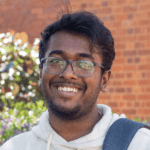 A student stands smiling in front of a garden with a backpack over his shoulder