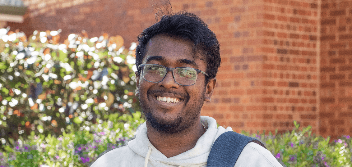 A student stands smiling in front of a garden with a backpack over his shoulder