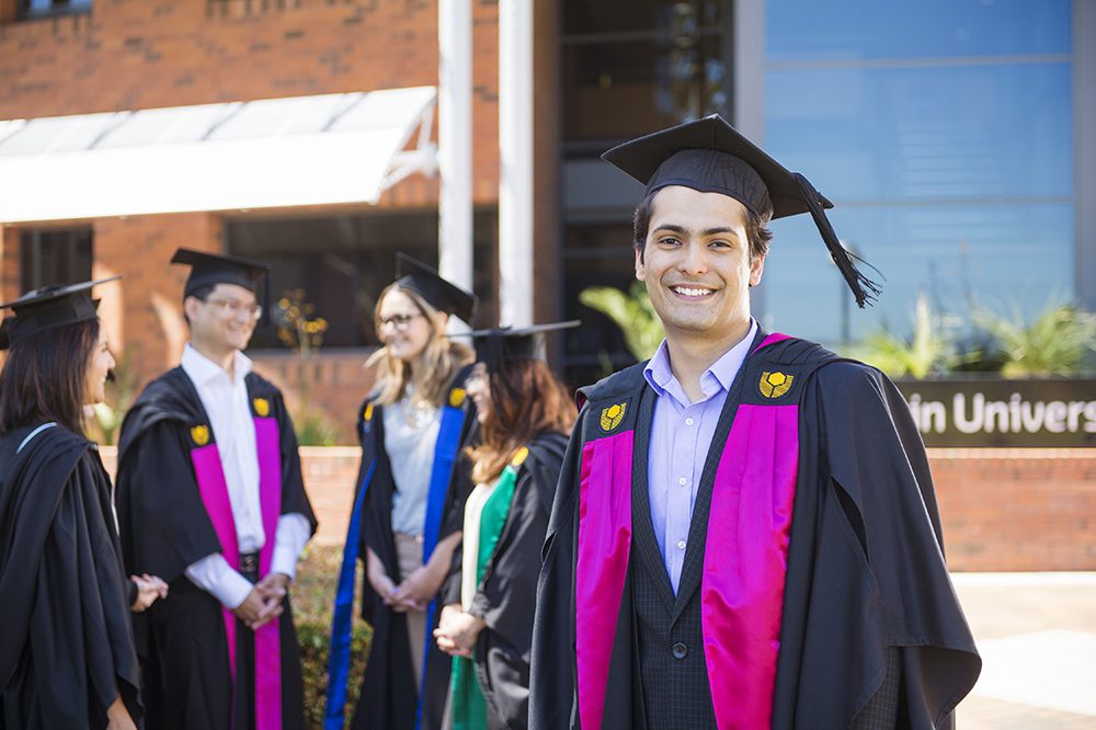 Young male graduate in graduation regalia and smiling and standing in front of a small group of graduates