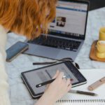 A student leaning over a laptop, iPad and notebook with pens and study accessories scattered around her.