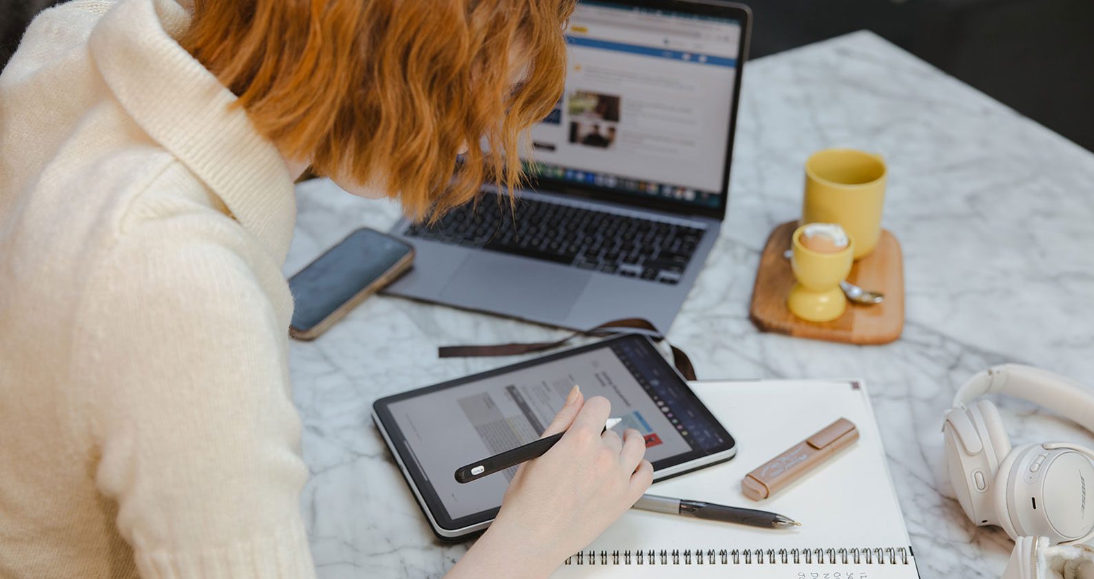 A student leaning over a laptop, iPad and notebook with pens and study accessories scattered around her.